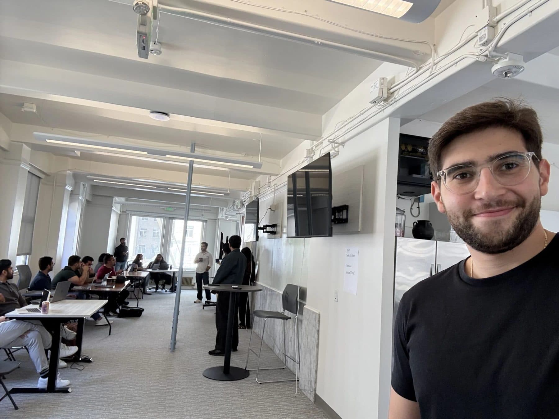 A selfie at the Emergent AI Hackathon, with teams working at laptops in the room behind